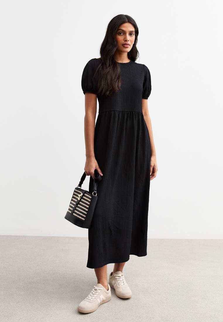 Abito midi nero a maniche corte con tessuto testurizzato e design a strati, abbinato a una borsa a tracolla a righe nera e crema e sneakers beige.