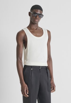 Joven con gafas de sol negras, camiseta blanca sin mangas y pantalones negros de talle alto, posando frente a un fondo gris liso.