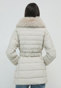 Giacca puffer beige con colletto in pelliccia, vita stretta, texture trapuntata e maniche lunghe. La vista posteriore mette in evidenza il materiale morbido e la forma.