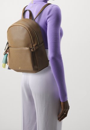 Personne portant un sac à dos en cuir marron perforé avec des pompons colorés, un haut à manches longues violet et un pantalon blanc, vue de dos.