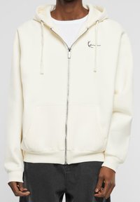 Sudadera cremosa con cremallera y cordones, bolsillos frontales, puños y dobladillo acanalados. Presenta un pequeño logo negro en el lado izquierdo del pecho. Textura de tela suave.