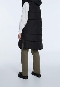 Zwarte donzen bodywarmer met capuchon, voorzien van een doorgestikt ontwerp, zijritsen en een rechte pasvorm, gecombineerd met olijfgroene broek en zwarte laarzen.