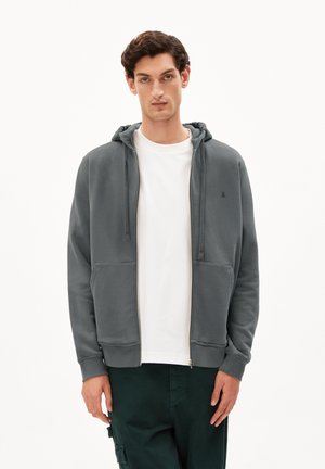 Grijze zip-up hoodie met een capuchon met trekkoord, voork officieel zorgvergoeding, en geribde manchetten. Draagt over een witte shirt, gecombineerd met donkergroene broeken.