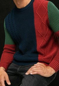 Maglione lavorato a maglia con una combinazione di sezioni blu scuro, rosse e verdi, caratterizzato da motivi testurizzati inclusi design a trecce e a costine.