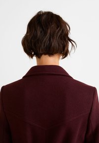 Personne aux cheveux bruns courts et ondulés portant un manteau en laine bordeaux foncé, vue de dos sur un fond blanc uni.