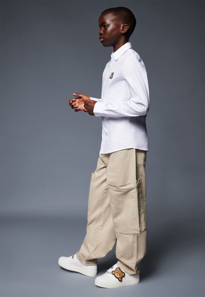 Camicia bianca button-up, pantaloni cargo beige con grandi tasche e scarpe slip-on bianche decorate con un design di orso. Stile semplice e funzionale.