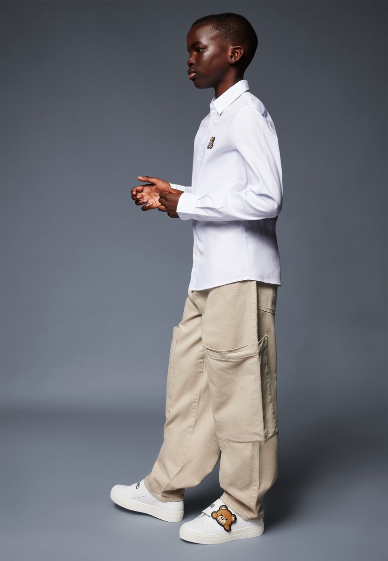 Camicia bianca button-up, pantaloni cargo beige con grandi tasche e scarpe slip-on bianche decorate con un design di orso. Stile semplice e funzionale.