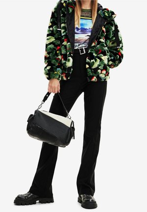 Donna che indossa una giacca di pelliccia ecologica verde con motivo, maglietta grafica a maniche lunghe, pantaloni neri, scarpe nere robuste, che tiene una borsa a mano bianca e nera.