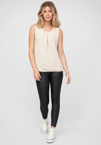 Beiger ärmelloser Oberteil mit Faltendetail, kombiniert mit schwarzen Leggings und weißen Turnschuhen. Glatte Textur; figurbetontes Design.