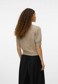 Maglione a maniche corte beige in maglia con vita aderente e tessuto texturizzato, abbinato a una lunga gonna nera. La vista posteriore mostra i capelli ricci.