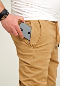 Beige Hosen mit einer Reißverschlusstasche halten ein graues Smartphone. Der Stoff hat eine weiche Textur. Ein Handgelenk mit schwarzen Armbändern ist sichtbar.