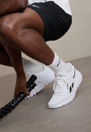 Λευκά αθλητικά παπούτσια με μαύρες λεπτομέρειες, διχτυωτό επάνω μέρος και μαξιλαρωμένο κολάρο. Φορεμένα με μαύρες βερμούδες και άσπρες κάλτσες, κρατώντας μια χοντρή μαύρη σχοινί.