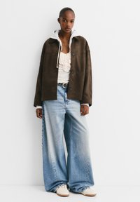 Veste en suédine marron superposée sur un haut crème à volants, accompagnée de jeans en denim bleu surdimensionnés. Des baskets blanches complètent le look.