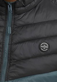 Černá prošívaná bunda s předním zipem, akcenty v barvě modrozelené a kulatou nášivkou s nápisem "Jack & Jones" na hrudi. Texturovaná látka.