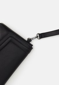 Pochette en cuir noir avec une surface texturée, dotée d'une sangle détachable et de quincaillerie en métal. Le design comprend des coutures discrètes et une forme élégante.