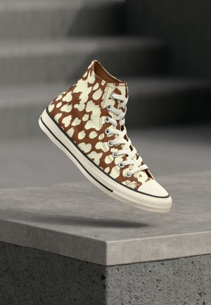 Καφέ και μπεζ αθλητικό παπούτσι τύπου leopard με κορδόνια σε λευκό χρώμα, αιωρούμενο πάνω από ένα τσιμεντένιο περίβολο με σκάλες στο φόντο.