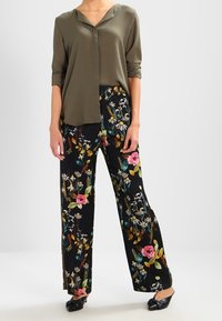 Blouse vert olive à coupe décontractée avec un col pointu, associée à un pantalon noir à jambes larges orné d'un imprimé floral coloré.