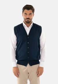Gilet lavorato a maglia blu navy con scollo a V e sei bottoni, indossato sopra una camicia bianca. Presenta orli a coste e giromanica per un look aderente.