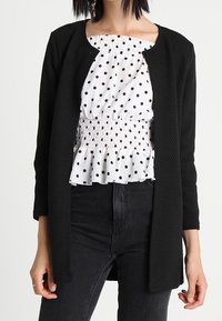 Svart texturerad kofta över en vit blus med svarta prickar, med smockdetaljer och en volangkant, parat med mörka jeans.