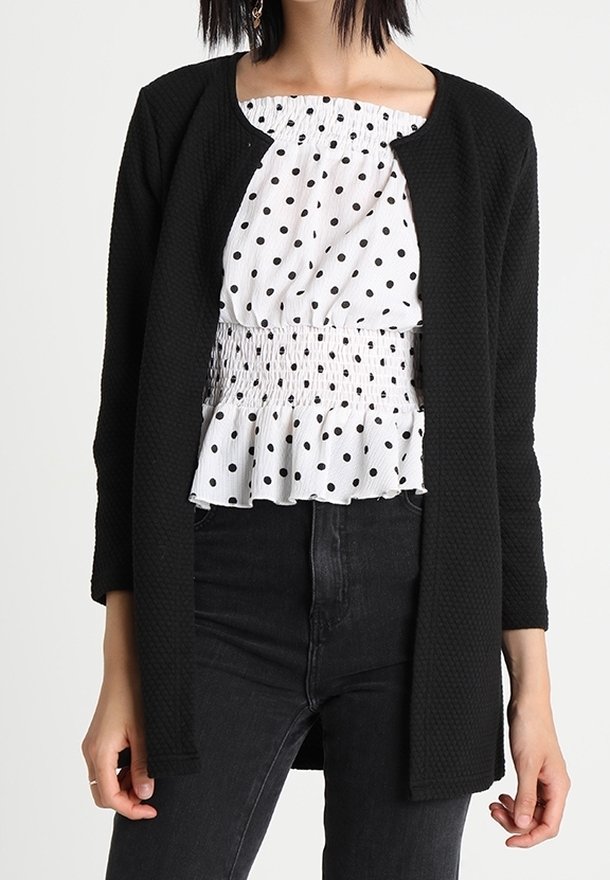 Svart texturerad kofta över en vit blus med svarta prickar, med smockdetaljer och en volangkant, parat med mörka jeans.