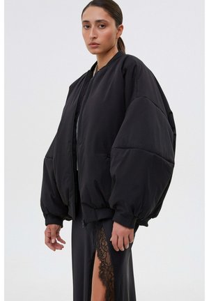 Kobieta ubrana w oversize'ową czarną kurtkę bomberkę i czarną spódnicę z wysokim koronkowym rozcięciem, stojąca na tle jednolitego białego tła.