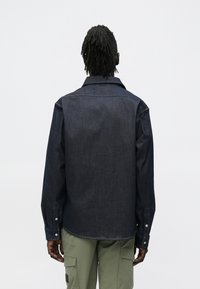 Mężczyzna widziany od tyłu, ubrany w ciemną kurtkę dżinsową i jasne zielone spodnie bojówki na jednolitym tle.