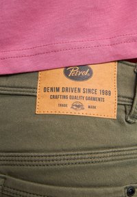 Pantalones de tela verde con una etiqueta de cuero marrón grabada con "Petrol" y texto, que presentan detalles de costura y una cintura visible.