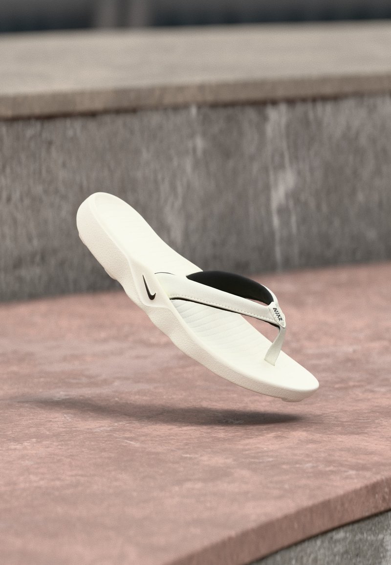 Λευκή σαγιονάρα Nike με μαύρη εσωτερική σόλα, στον αέρα πάνω από ροζ αποχρωματισμένο έδαφος και φόντο γκρι πέτρας.