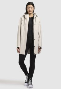 Veste imperméable beige avec capuche, boutons à l'avant, accents noirs et manches longues, portée par-dessus des leggings de sport noirs et des baskets montantes.