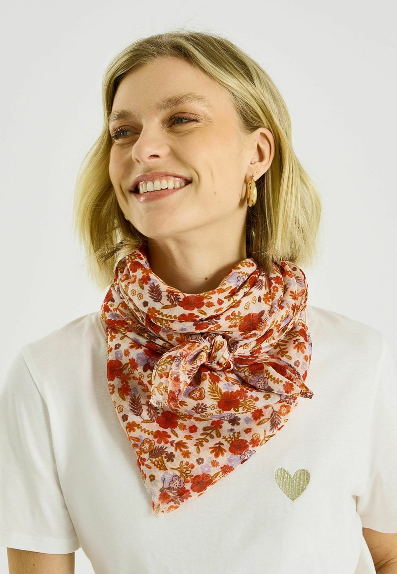 Mujer sonriente con cabello corto y rubio, lleva pendientes de aro dorados, una camisa blanca con un pequeño corazón dorado y una bufanda floral naranja anudada alrededor del cuello.