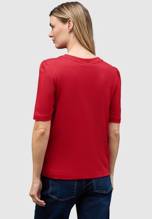 T-shirt rouge à manches courtes avec un décolleté arrondi et de légères manches bouffantes. Le matériau semble doux avec une texture lisse. Porté avec un jean bleu foncé.
