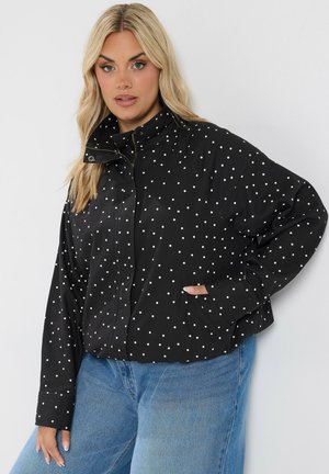 Mujer rubia que lleva una chaqueta negra con lunares blancos y vaqueros azules, de pie con una mano en el bolsillo frente a un fondo simple.