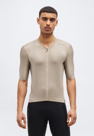 Moški, ki nosi bež kolesarski dres s kratkimi rokavi z zadrgo in črne kolesarske hlače, stoji ob preprostem svetlem ozadju.