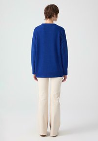 En person med kort brunt hår bär en lös långärmad tröja i kungligt blått och vida byxor i off-white, stående mot en enkel ljus bakgrund.