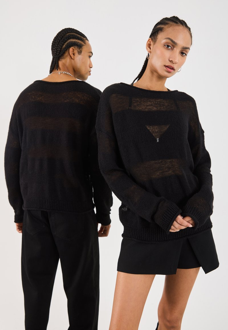 Schwarzer Strickpullover mit lockerer Passform, der transparente horizontale Streifen und gerippte Bündchen aufweist. Kombiniert mit schwarzen Hosen und Shorts.
