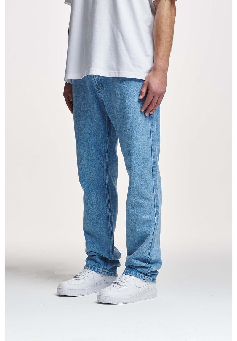 Homme portant un jean bleu clair, des baskets blanches et un t-shirt blanc, debout devant un fond uni clair.