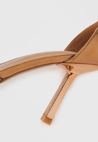 Smeđe kožne sandale s visokim potpeticama i sjajnom ružičastom zlatnom stiletto potpeticom. Ima široku traku i minimalistički dizajn. Glatka tekstura.