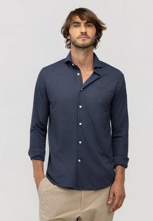 Hombre con cabello castaño que lleva una camisa azul marino de manga larga con botones blancos y pantalones beige, de pie con las manos en los bolsillos frente a un fondo claro.