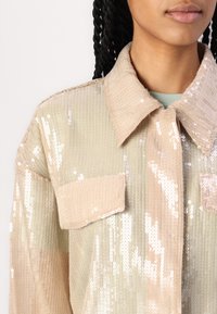 Beige sequined shirt jacket with a textured finish, featuring a classic collar and two flap pockets. Shiny, reflective surface.