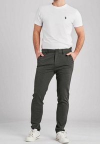 Hombre vestido con camiseta blanca de manga corta con logo, pantalones oscuros slim-fit y zapatillas blancas, de pie con las manos en los bolsillos.