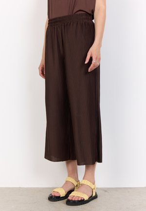 Brown, pleated culottes with an elastic waistband, paired with yellow frilled sandals and a textured surface on a neutral background.