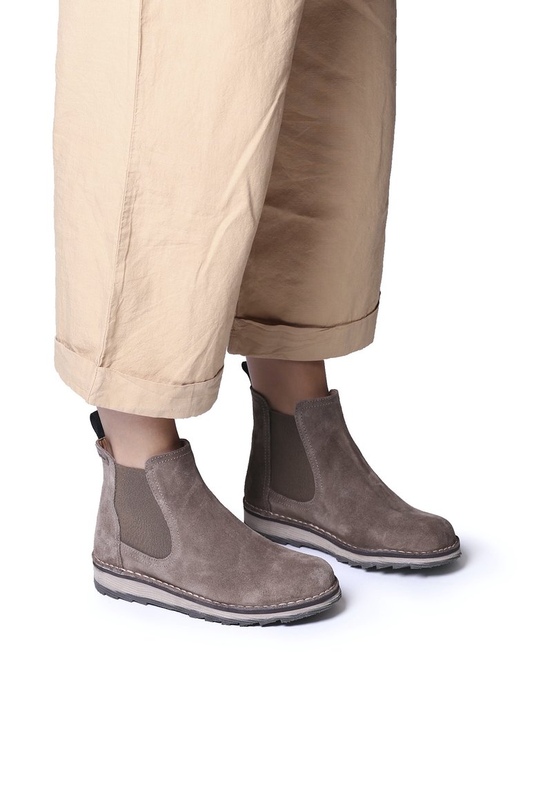 Bottes Chelsea en daim taupe montant à la cheville portées avec un pantalon beige raccourci sur un fond blanc.