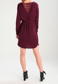 Burgundy klänning med långa ärmar, texturerad spetsbak, åtstramad midja och en lätt utsvängd nederdel. I kombination med svarta ankelskor.