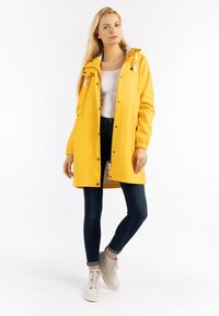 Cappotto impermeabile giallo con cappuccio, dotato di polsini elasticizzati e bottoni a pressione. Abbinato a jeans scuri e sneakers di colore chiaro.