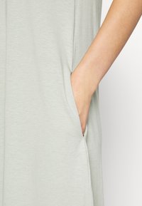 Peça de vestuário cinza claro com uma textura suave, apresentando um bolso lateral e um corte descontraído. O braço revela um tom de pele suave contra o tecido.