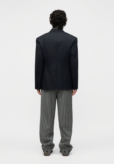 Άτομο με σγουρά μαύρα μαλλιά, φορώντας μαύρο σακάκι και φαρδιά γκρι παντελόνια με κάθετες λευκές ρίγες, στέκεται με την πλάτη σε λευκό φόντο.