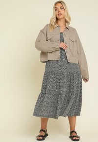 Ljusbrun, kort jacka med framfickor, bärs över en svart blommig, lager-på-lager maxi-klänning. Svarta sandaler fulländar outfiten.