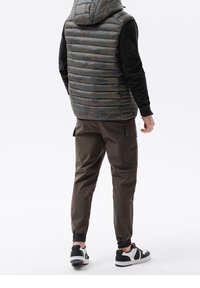 Kapuzensack, ärmellose Steppweste im Camouflage-Muster, kombiniert mit braunen, schmal geschnittenen Cargo-Hosen und schwarz-weißen Sport-Schuhen.