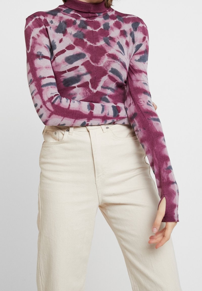 Personne portant un haut à manches longues tie-dye violet et gris avec des trous pour les pouces et un pantalon taille haute écru sur un fond uni.