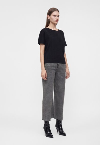 Femme debout portant un haut noir à manches courtes, un jean gris taille haute à jambes larges et des bottines noires à talons sur fond blanc.
